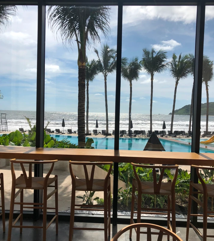 Terraza con sillas y piscina, palmeras y mar bajo cielo nublado.