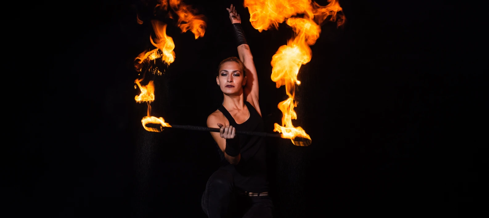 Mujer con bast&oacute;n de fuego, levanta la mano con llamas naranjas al fondo.