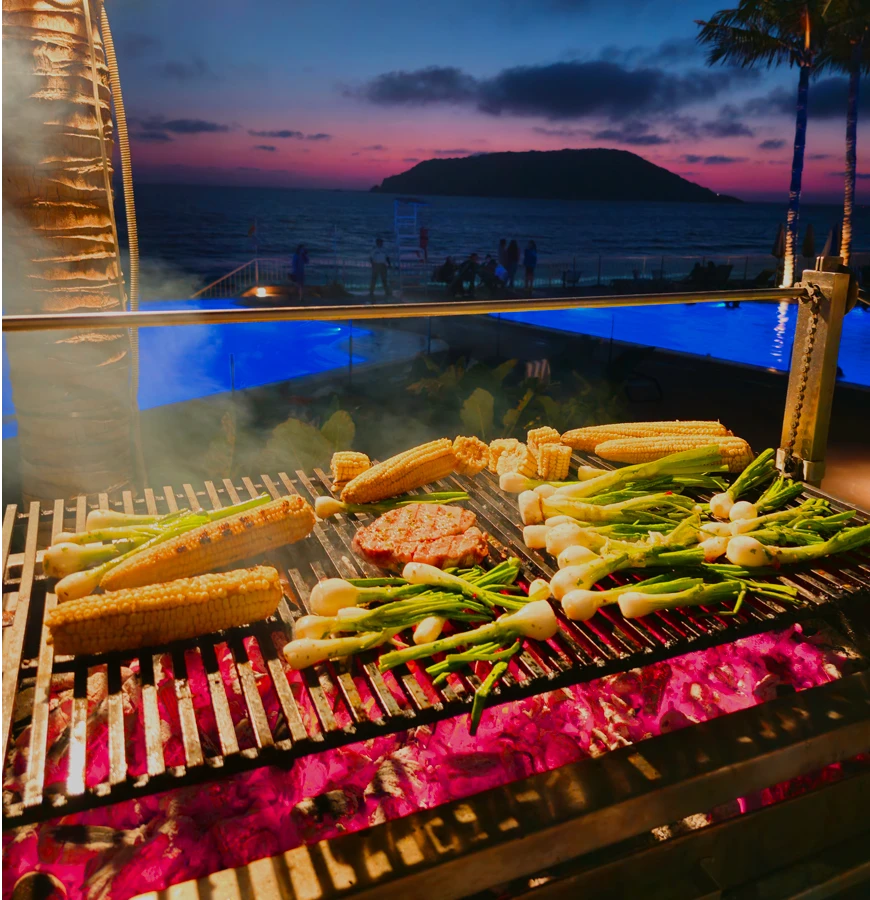 Parrilla con ma&iacute;z, verduras y atardecer frente al mar, junto a piscina.