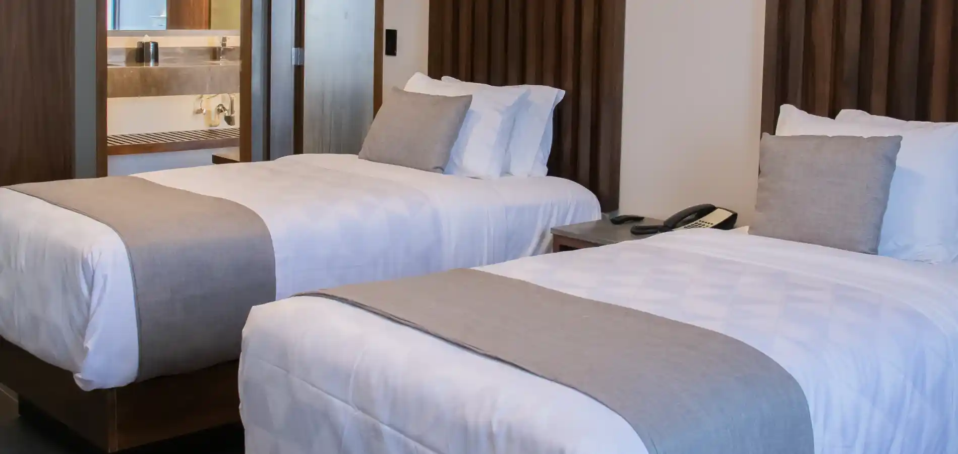 Habitaci&oacute;n de hotel con tres camas y almohadas claras, escritorio y puerta al ba&ntilde;o