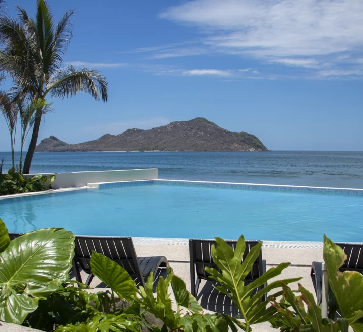 Piscina frente al mar con palmeras y una isla monta&ntilde;osa al fondo