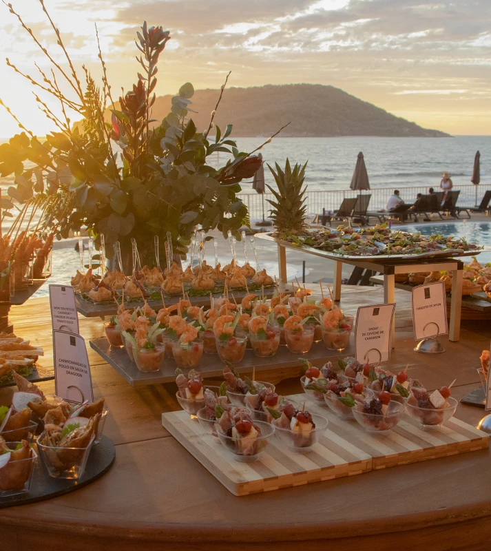 Mesa con canap&eacute;s y atardecer en la costa, mar y velas al fondo