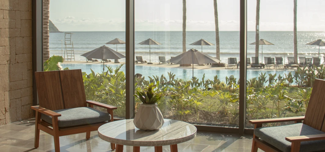Vista desde un hotel: terraza con sillones, mesa y palmeras junto al mar.