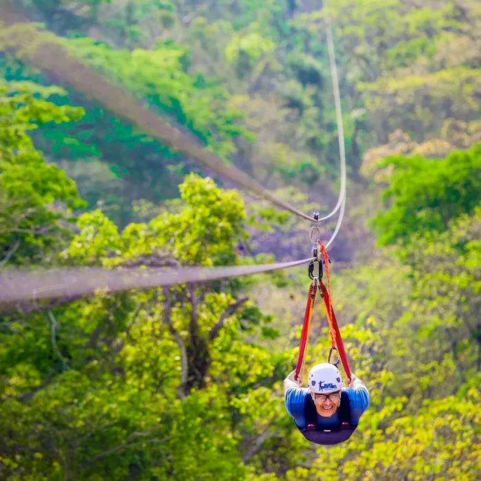Persona en tirolesa colgando sobre un bosque verde y monta&ntilde;as, con arn&eacute;s