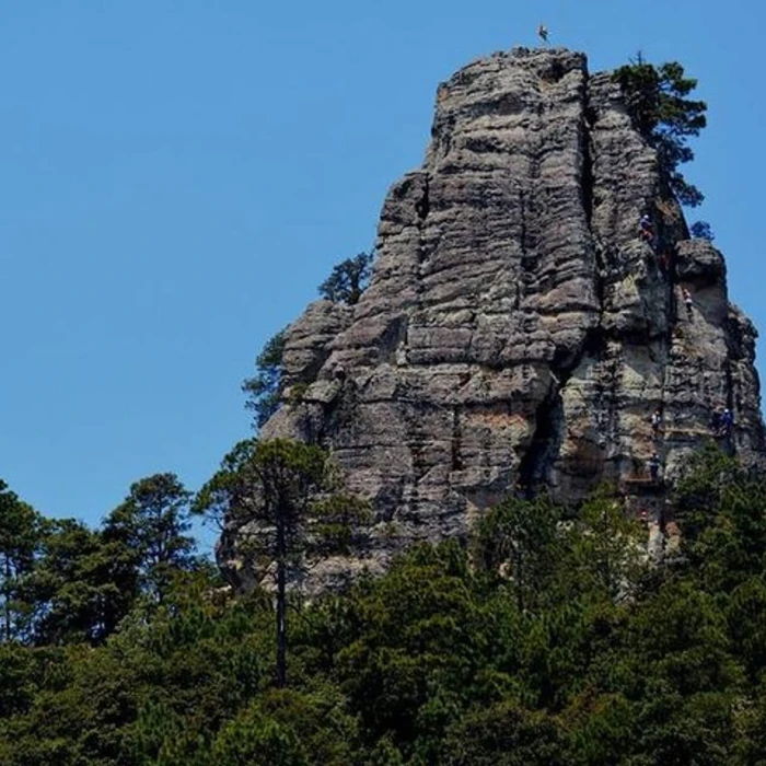 Roc&oacute;n rocoso con &aacute;rboles verdes y personas en la cima, cielo azul.