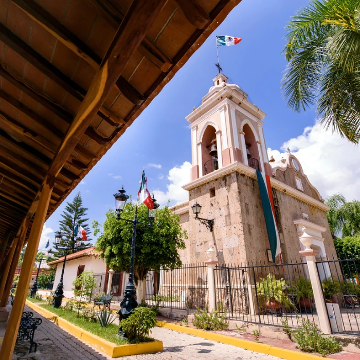 Torre blanca con campana, banderas y palmeras bajo un cielo azul