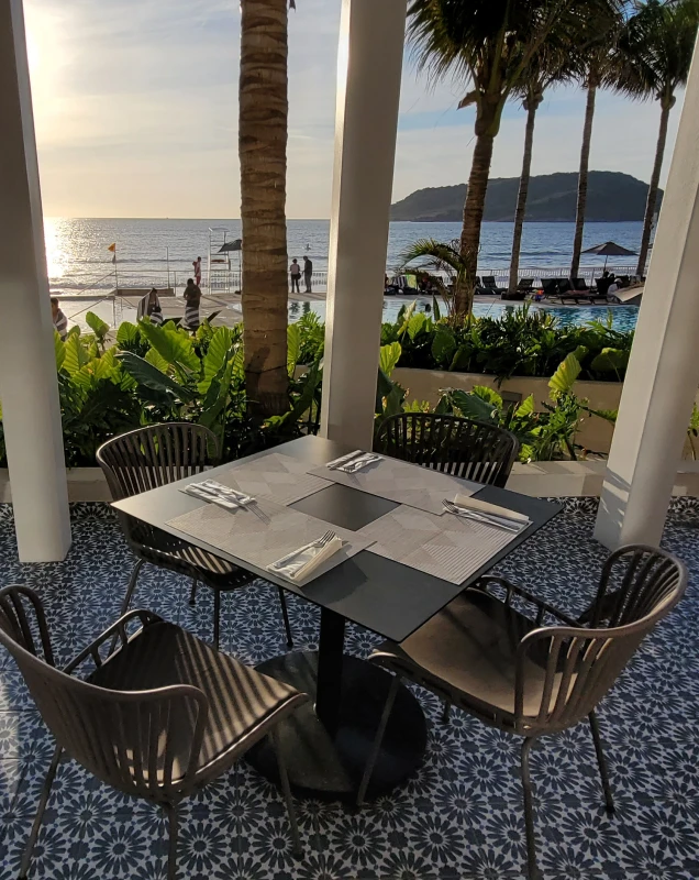 Mesa servida en terraza con palmeras y vista al mar al atardecer.