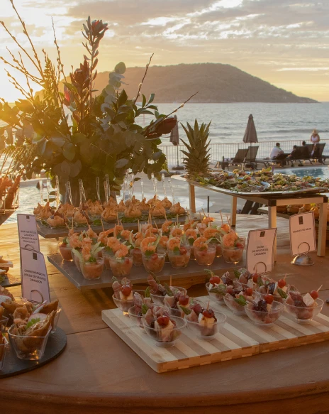 Buffet con canap&eacute;s y mariscos al atardecer, junto al mar y mesas al aire libre.