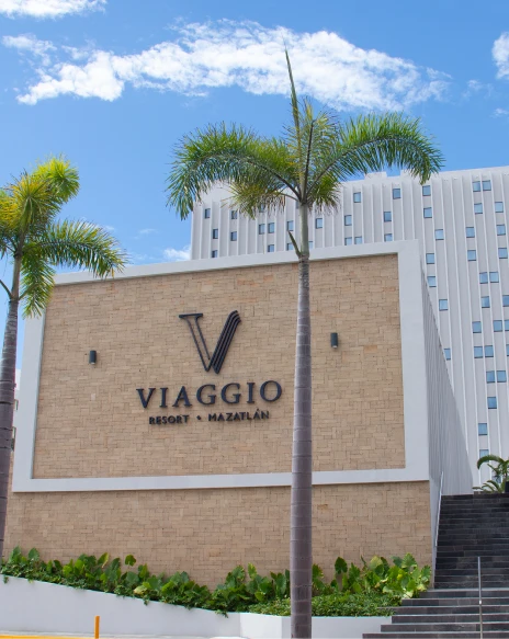 Cartel de &ldquo;Viaggio Resort Mazatl&aacute;n&rdquo; con palmeras y edificio bajo cielo azul.