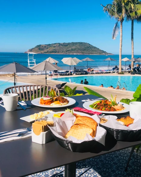 Mesa con platos y comida frente a piscina y mar azul, palmeras al fondo.