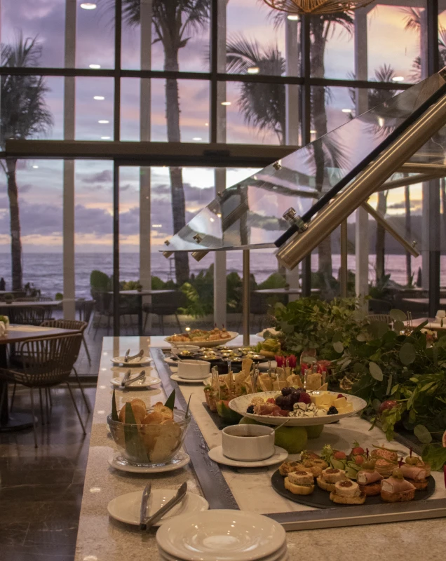 Desayuno en buffet junto al mar, con mesas y plantas al atardecer.