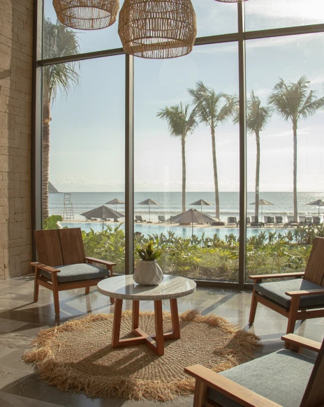 Terraza con sillas de madera, mesa y vista al mar con palmeras.