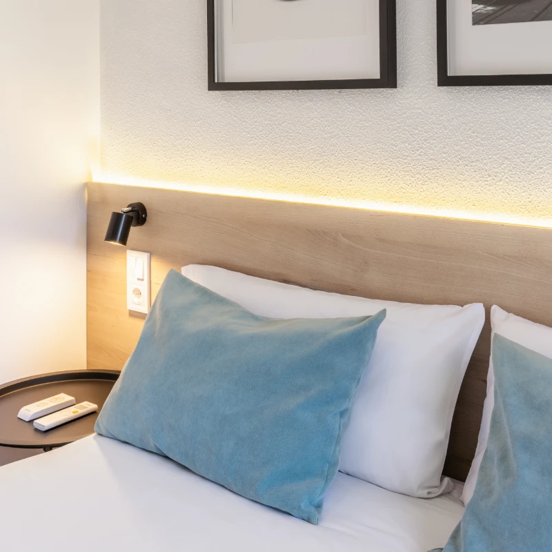Cama con almohadas azules y blancas, l&aacute;mpara y pared con luz c&aacute;lida