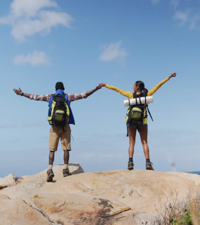 Dos excursionistas con mochilas, en una roca, con los brazos al cielo