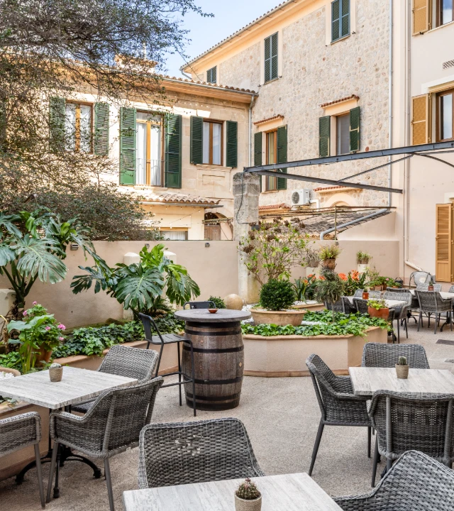 Patio con mesas, sillas y plantas frente a una casa de piedra con persianas