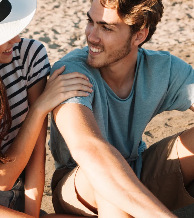 Joven con gorra azul sonr&iacute;e abrazando a un ni&ntilde;o en la playa.
