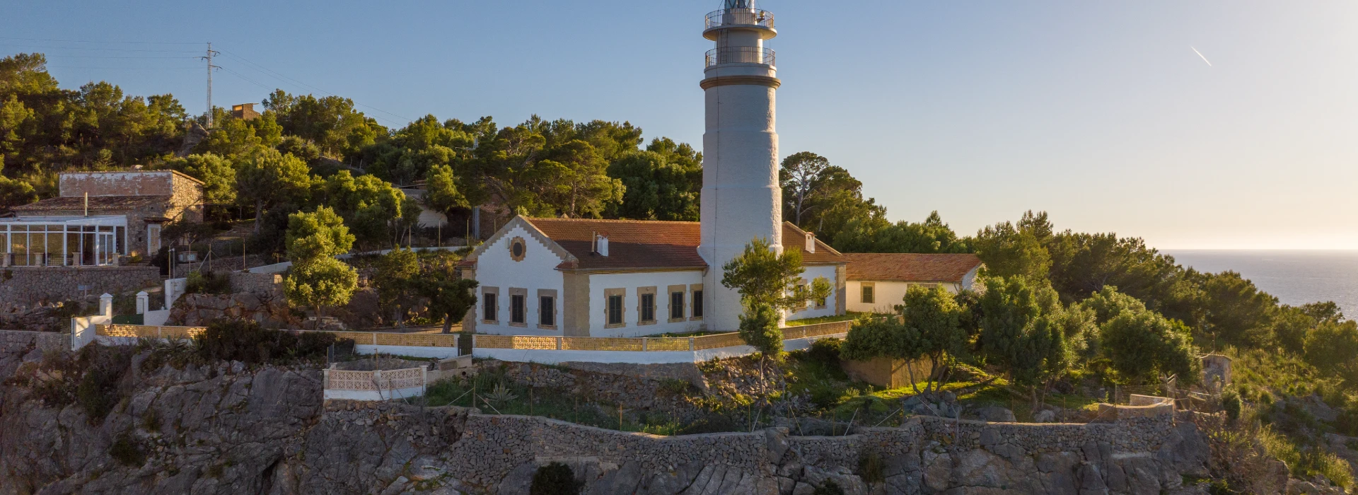 Faro blanco junto a casas y vegetación, al atardecer junto al mar