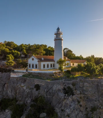 Faro blanco sobre acantilado, con cielo azul y vegetación alrededor.