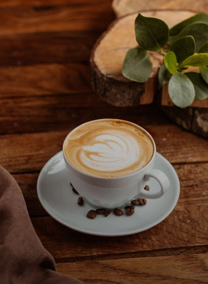 Taza de caf&eacute; con latte art sobre mesa de madera, con hojas verdes al fondo.