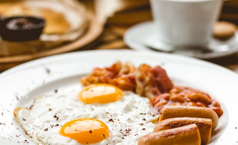 Plato de huevos fritos con tocino y salchicha, desayuno sobre mesa r&uacute;stica.