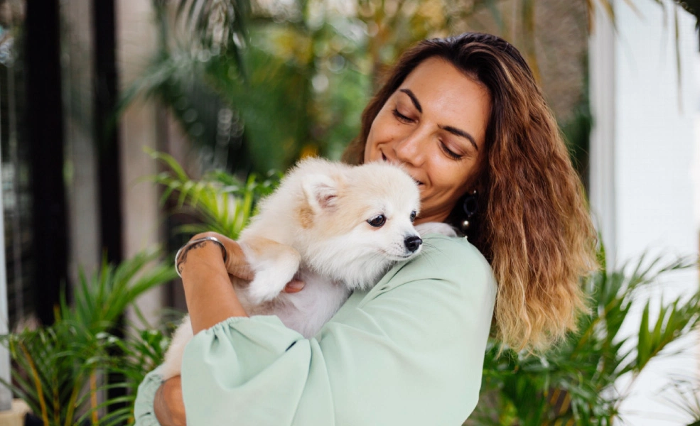 Mujer abraza a un cachorro blanco en un jard&iacute;n, con ojos cerrados.