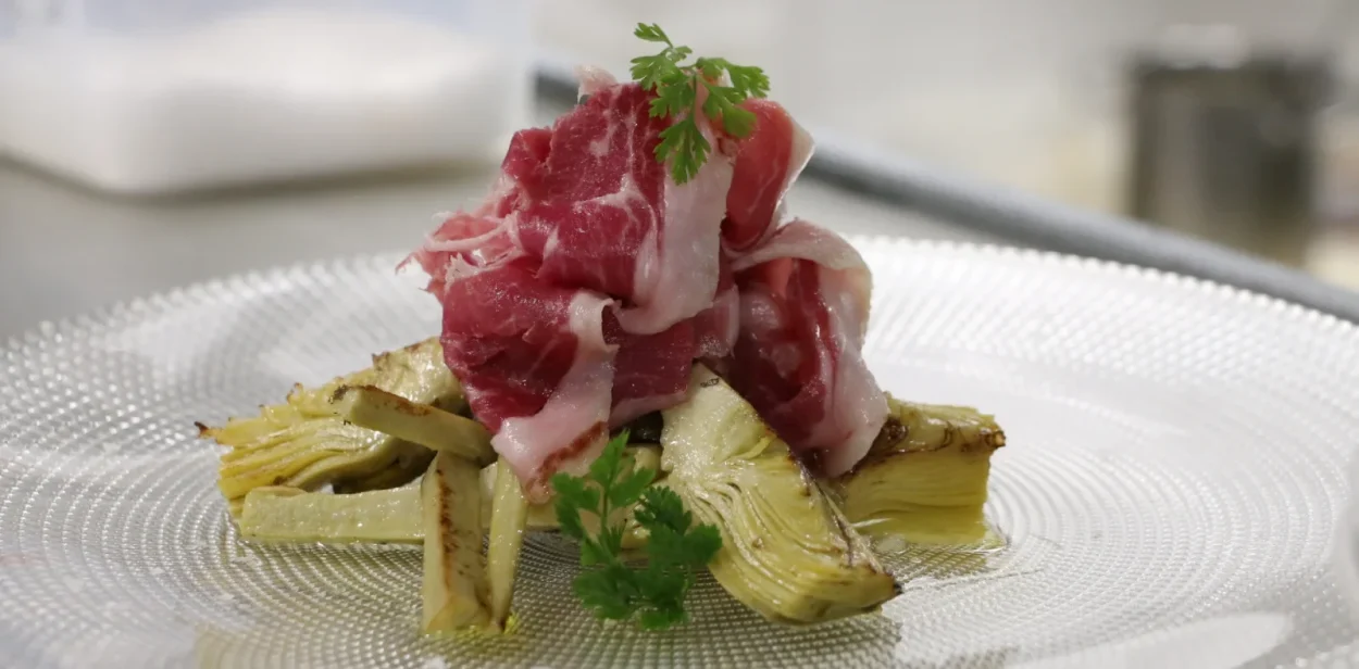 Jam&oacute;n ib&eacute;rico sobre alcachofas con perejil fresco, en plato blanco.
