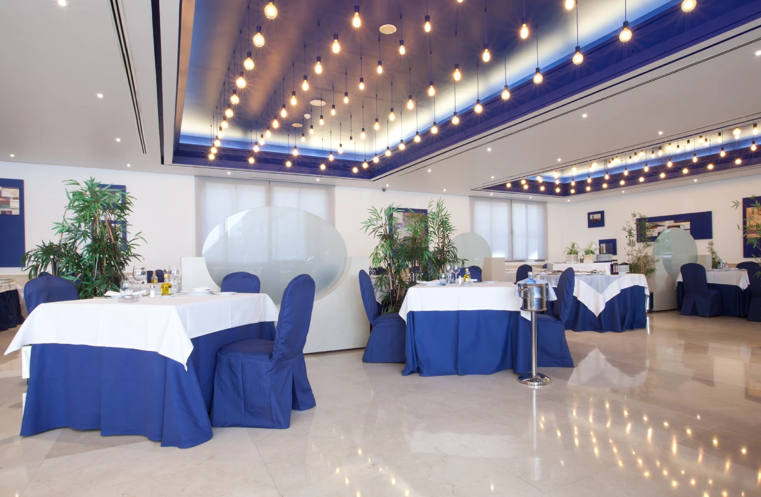 Sal&oacute;n de banquete con mesas blancas y manteler&iacute;a azul, luces colgantes y bamb&uacute;