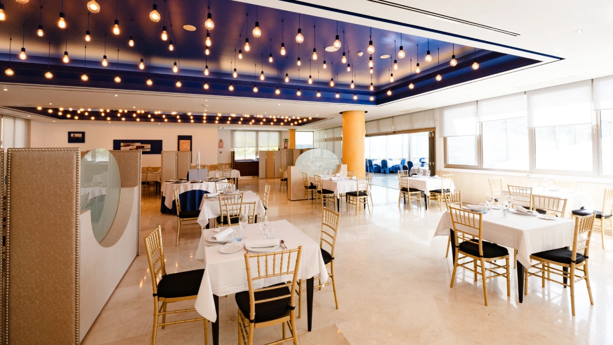 Sal&oacute;n de eventos con mesas vestidas, sillas doradas y luces colgantes.