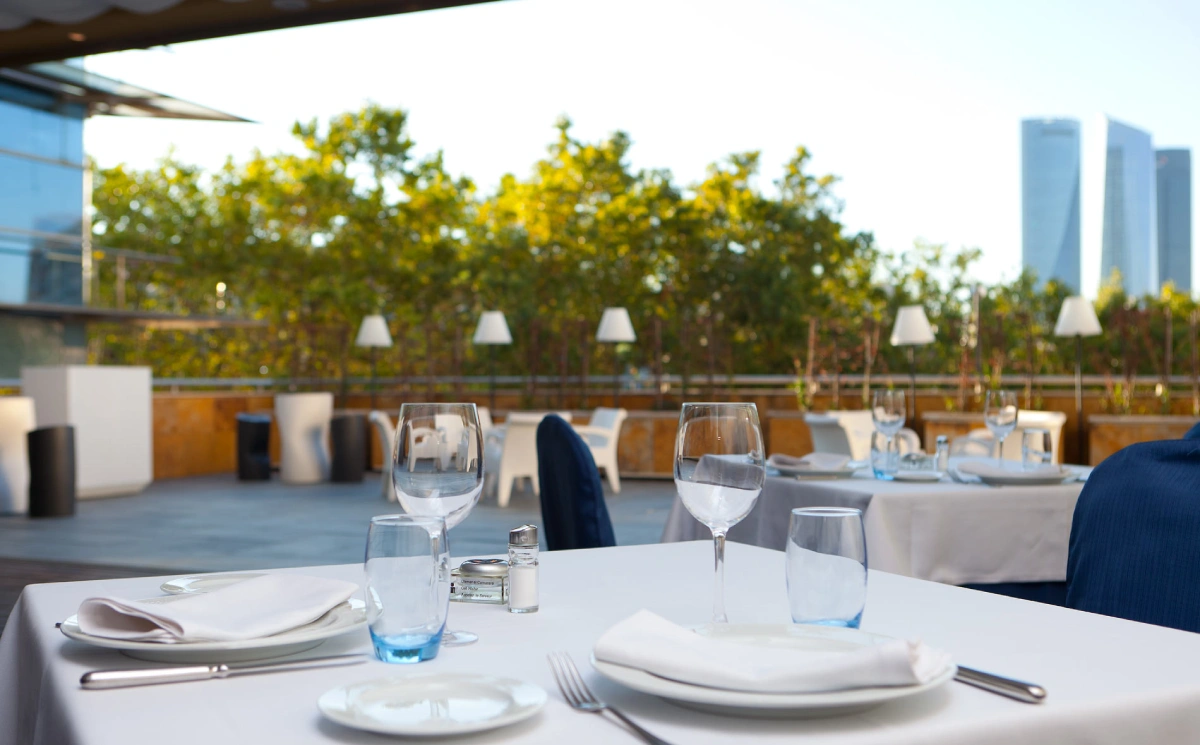 Mesa puesta al aire libre con copas y manteles, terraza con l&aacute;mparas y &aacute;rboles.