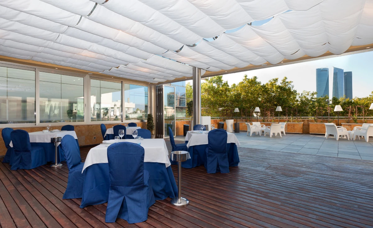 Terraza con mesas cubiertas y sillas azules bajo carpas blancas, vista urbana al fondo