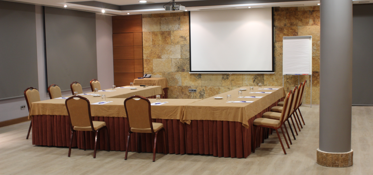Sala de reuniones con mesa larga, sillas y pantalla de proyecci&oacute;n.