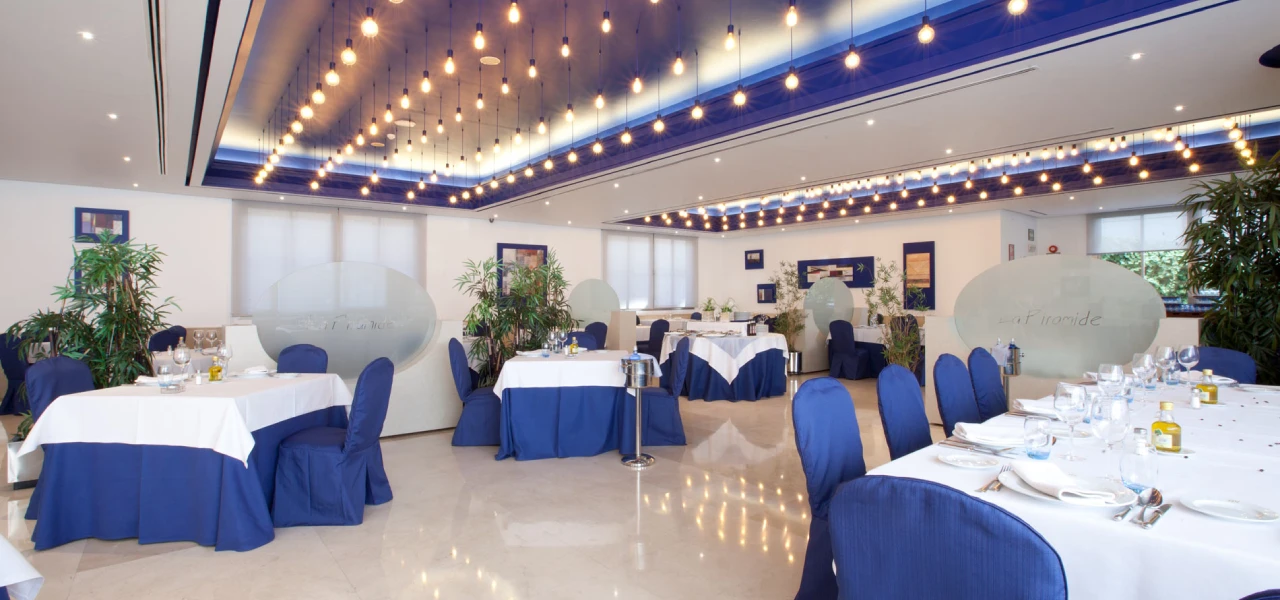 Sal&oacute;n de eventos con mesas, sillas azules y luces colgantes c&aacute;lidas.