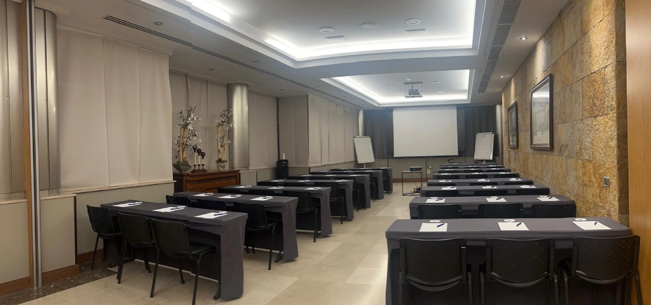 Sala de reuniones con mesas negras y proyector, iluminaci&oacute;n de techo y persianas