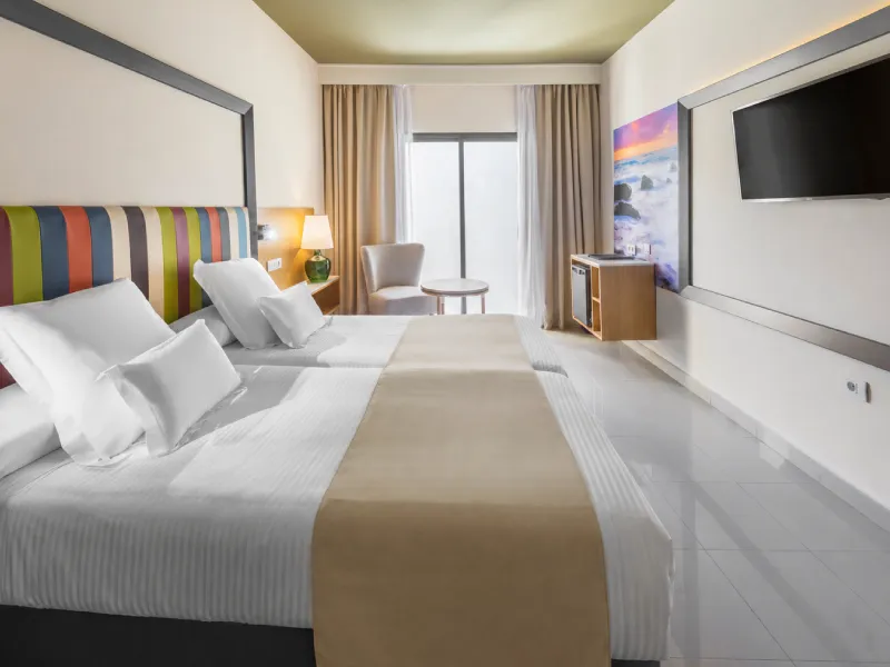 Habitaci&oacute;n de hotel moderna con cama doble, cortinas beige y TV.