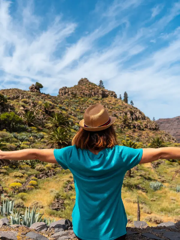 Mujer con sombrero azul, brazos abiertos, paisaje monta&ntilde;oso y cielo azul