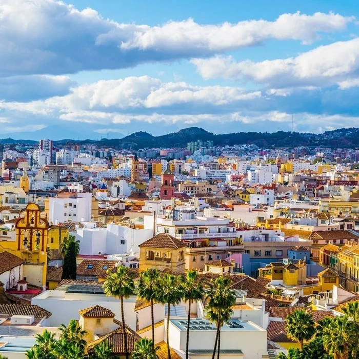 Panorámica urbana con tejados, palmeras y colinas bajo un cielo azul con nubes