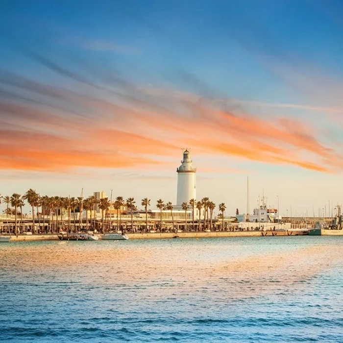 Faro blanco junto a palmeras y barcos en el atardecer.