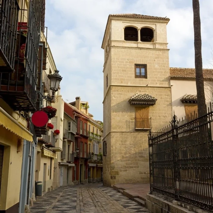 Calle estrecha con torre de iglesia de piedra y cielo nublado.