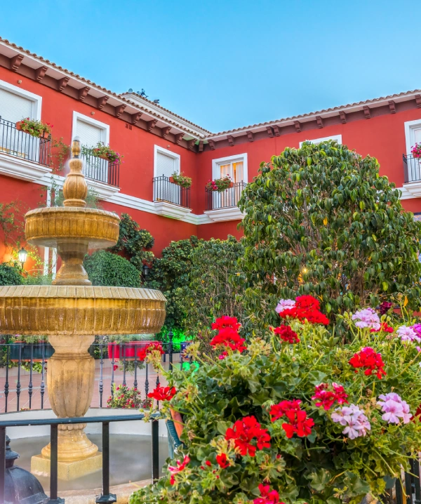 Patio andaluz con fuente dorada, flores rojas y rosadas y paredes rojas.