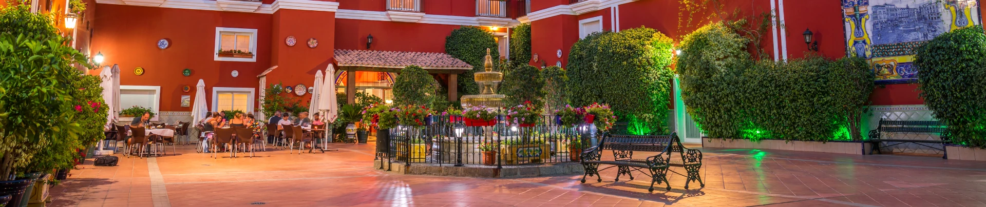 Patio con fuente y mesas bajo toldos, rodeado de muros rojos y plantas verdes.