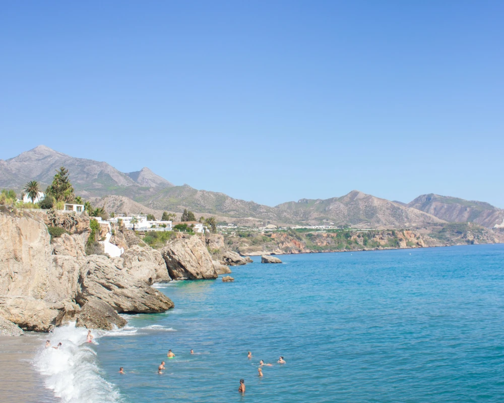 Playa rocosa con ba&ntilde;istas, acantilados y monta&ntilde;as al fondo, mar azul.