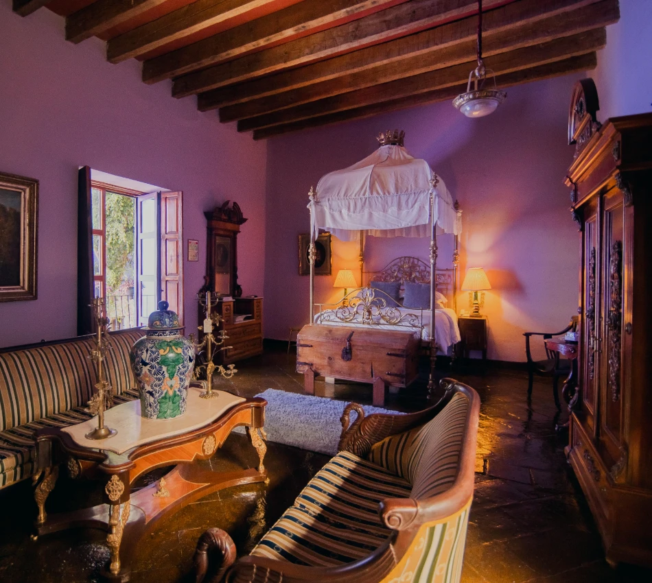 Interior de dormitorio colonial con cama con dosel, sillones y l&aacute;mparas encendidas