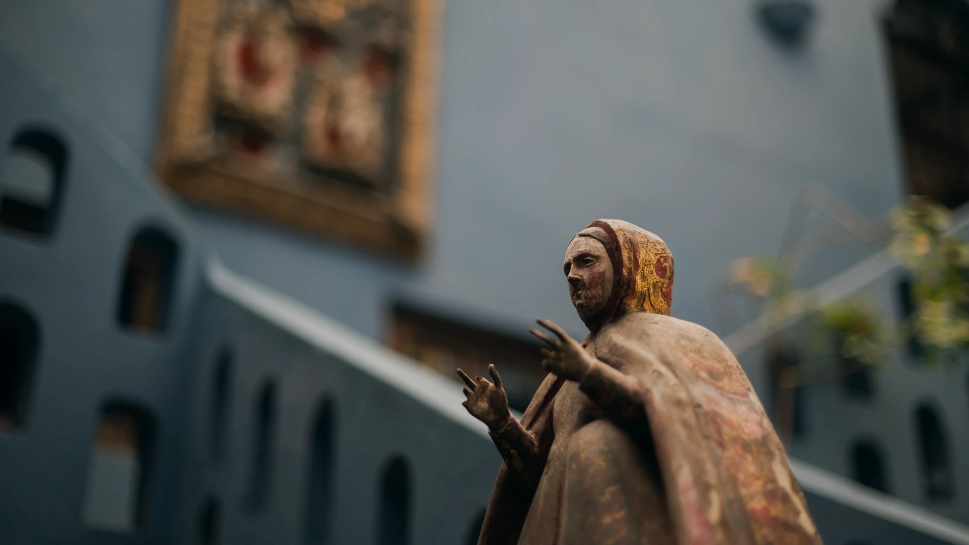 Estatua de un santo saludando en primer plano, con edificio al fondo (desenfocado)