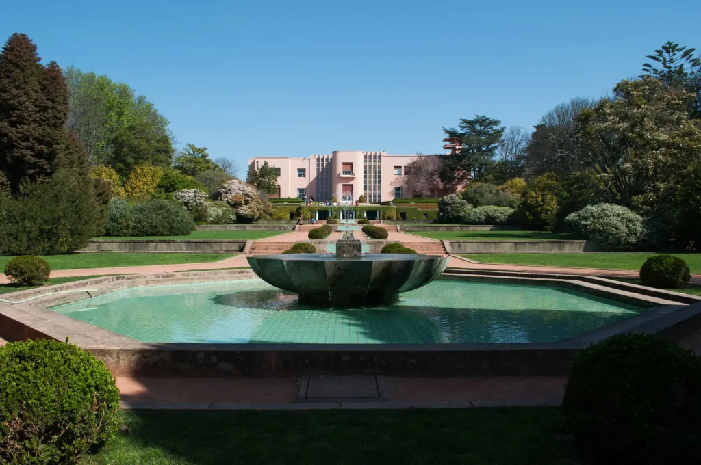 Jardim com fonte no centro e pr&eacute;dio ao fundo, c&eacute;u azul e &aacute;rvores