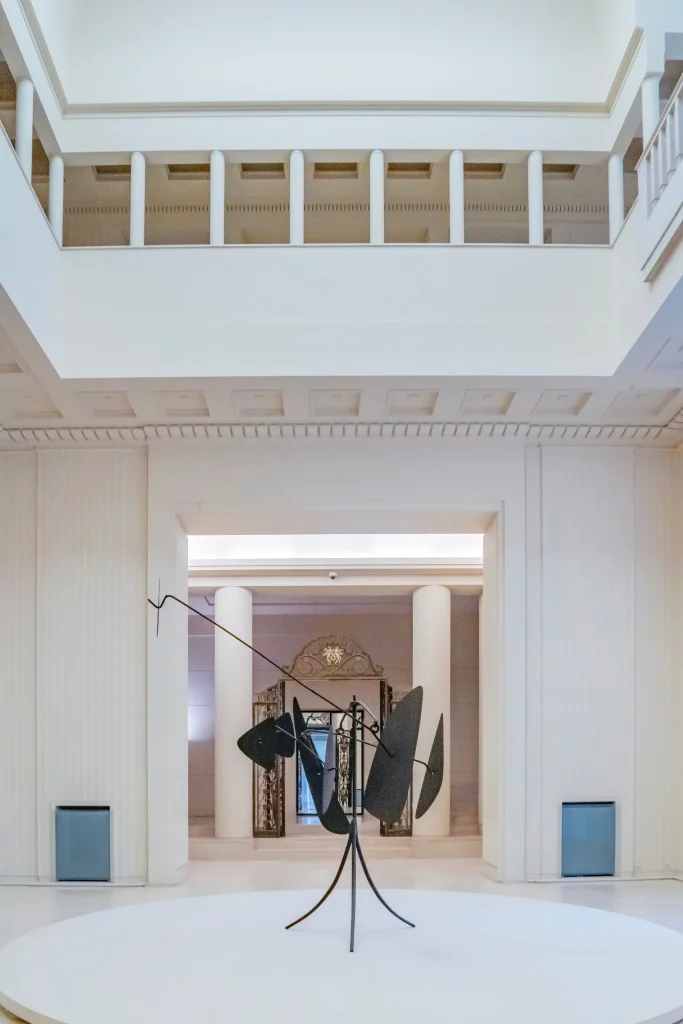 Vista interna com colunas brancas, c&uacute;pula clara e escultura met&aacute;lica no ch&atilde;o