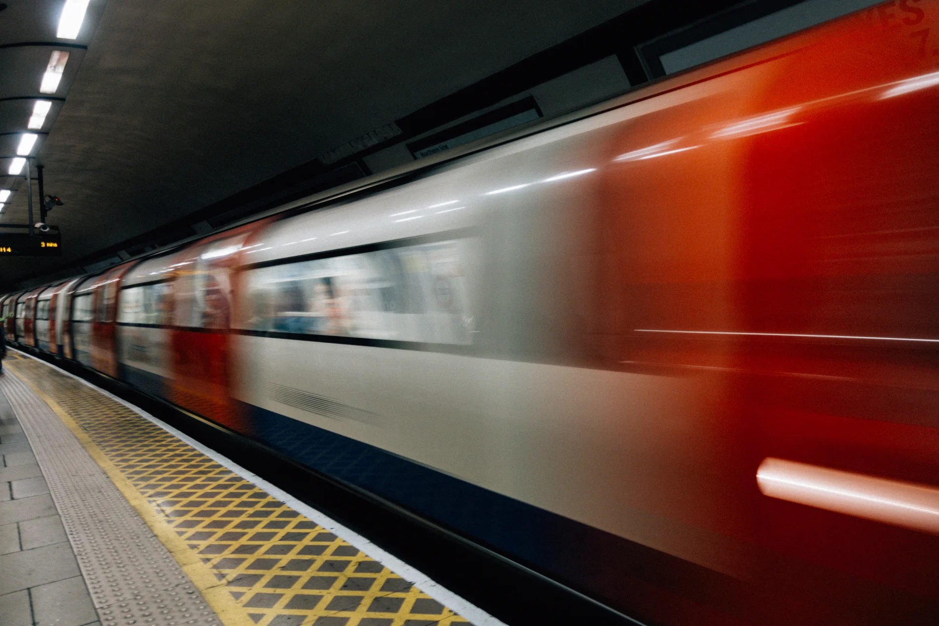 Trem laranja e branco passando pela plataforma da esta&ccedil;&atilde;o, com borr&atilde;o de movimento