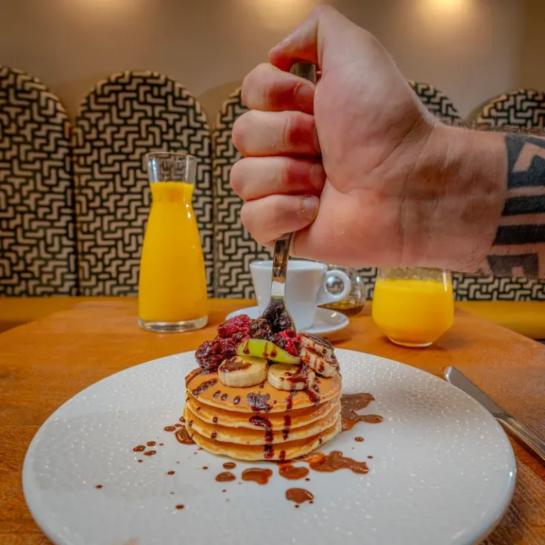 Mano sirve pancakes con frutas y jarabe en mesa con jugos naranjas