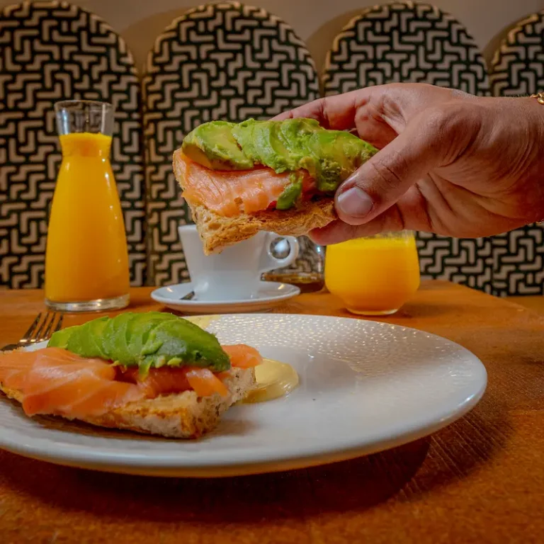 Mano sostiene un s&aacute;ndwich de salm&oacute;n y aguacate con fondo de mesa y jugos