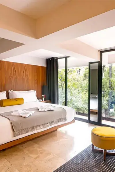 Dormitorio moderno con cama, ventanas de vidrio y sill&oacute;n amarillo.