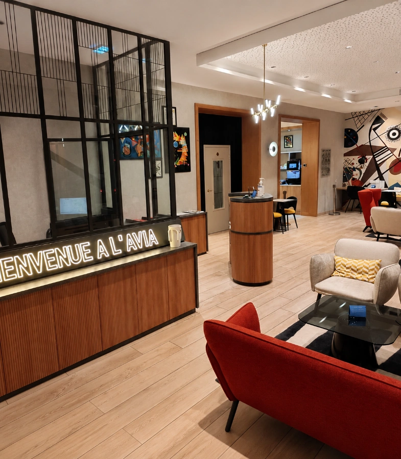 R&eacute;ception moderne d&rsquo;h&ocirc;tel, comptoir noir &ldquo;Bienvenue &agrave; l&rsquo;Avia&rdquo;, salon avec fauteuils rouges.